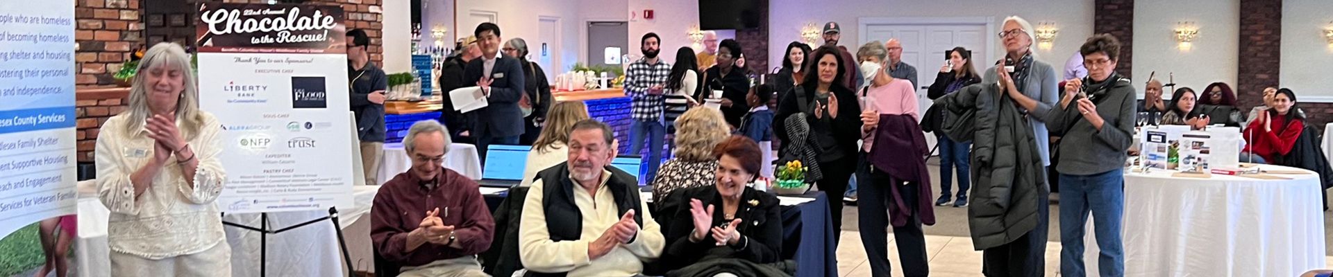 People applauding at an event. There is a sign that reads "22nd Annual Chocolate to the Rescue!" and has a list of sponsors below.