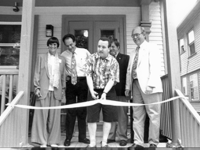 Group of people standing in front of a building entrance at an opening ceremony. A man in the middle is cutting a ribbon