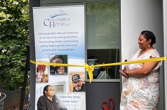 Woman cutting the ribbon at an opening ceremony. To her right is a sign with the Columbus house logo and text that reads "Serving people who are homeless or at risk of becoming homeless by providing shelter and housing and by fostering their personal growth and independence."