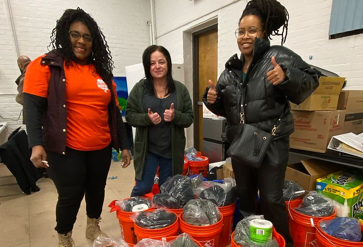 Three woman gathered around large orange buckets smiling with thumbs up
