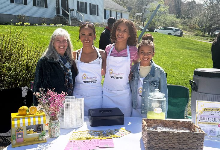 Three girls posing and smiling with an older woman at a lemonade stand fundraiser. The girls are wearing aprons that say "Penelope's Lemonade. Ending homelessness one cup at a time."